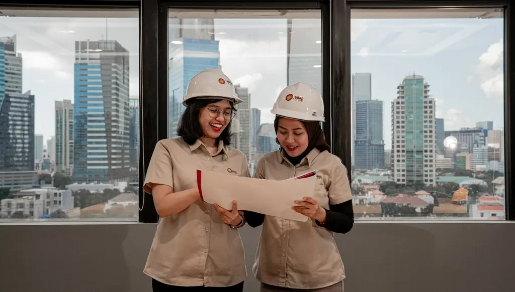 Two engineers in hard hats reviewing documents beside a window